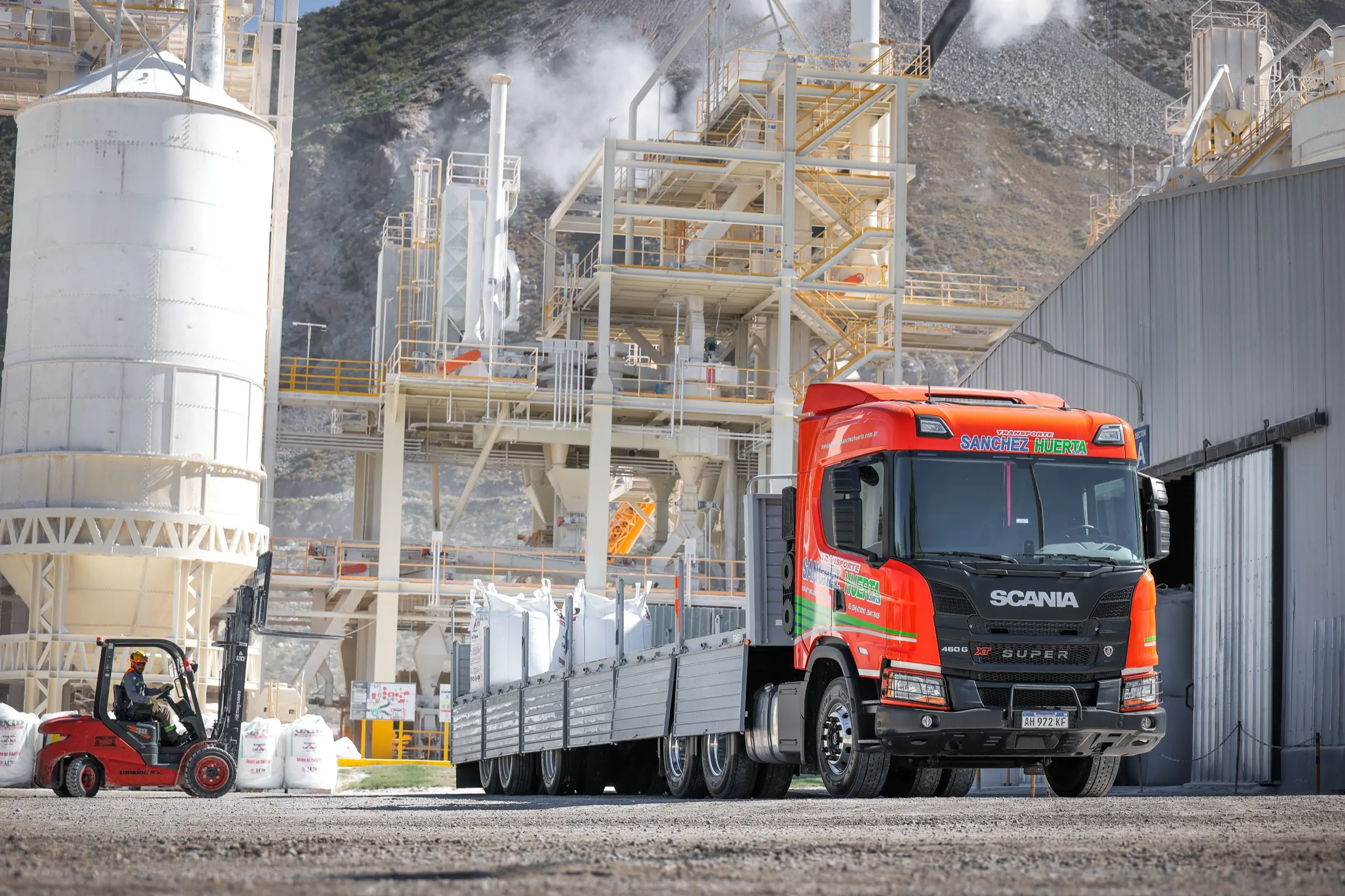 Camión Scania de Transporte Sanchez Huerta con acoplado de barandas cargado con bolsones blancos, y un autoelevador cargando más bolsones a un costado, en una planta industrial con silos y estructuras metálicas al pie de un cerro