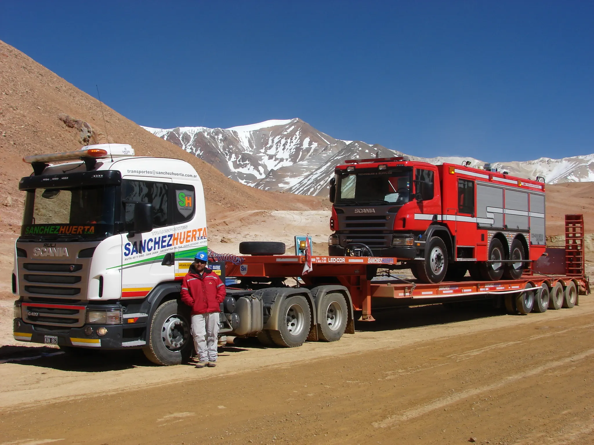 Camión con carretón de baja cama transportando un vehículo de emergencia en la operación minera de Lama, en alta cordillera, con un operario junto al equipo