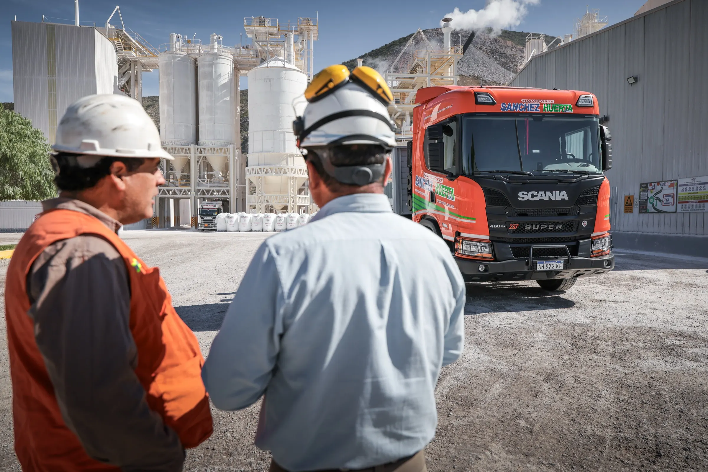 Dos operarios con casco y protectores auditivos conversando frente a un camión Scania, en una planta industrial con silos al pie de un cerro
